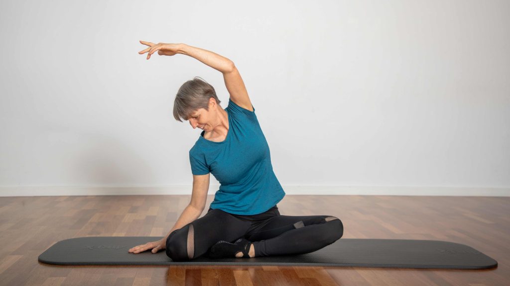 Frau sitzt im Schneidersitz und lehnt sich seitlich nach unten mit ausgestrecktem Arm.