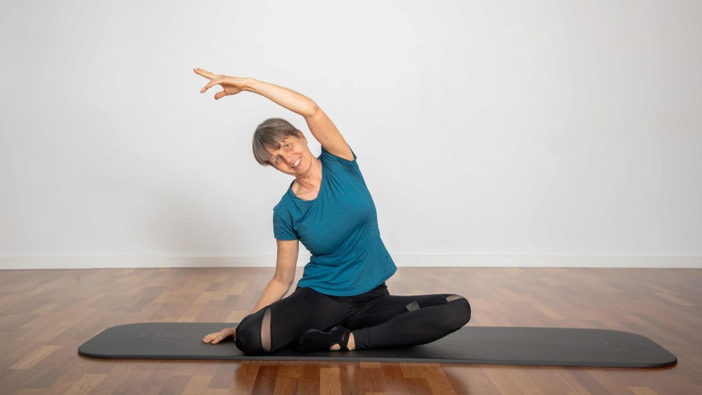 Frau sitzt auf einer Matte und streckt den Arm über den Kopf zur Seite.